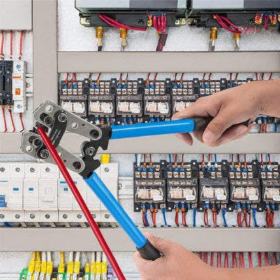 Mains tenant pince a sertir bleue avec cosse gaine à rouge devant tableau électrique